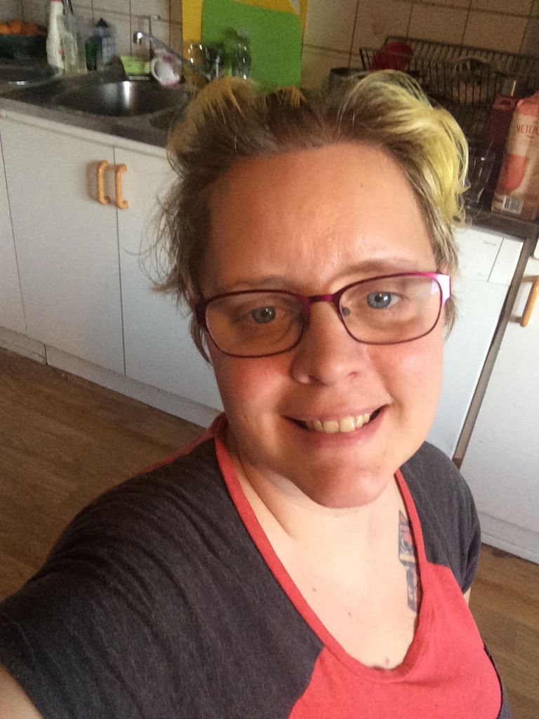 Lindha med blont hår och glasögon, står i ett kök.