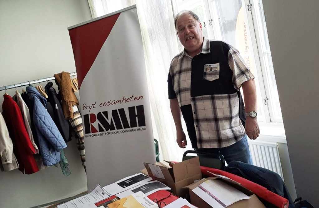 Stefan Wallerek iklädd rutig skjorta, stående vid ett bord med broschyrer från RSMH. 